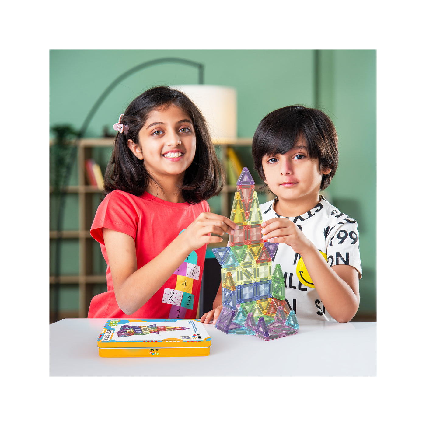 A smiling girl and a boy build a tall rocket tower together using colorful, translucent magnetic tile blocks.