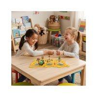 Two young girls laughing and playing with a dinosaur track puzzle toy on a wooden table in a playroom.