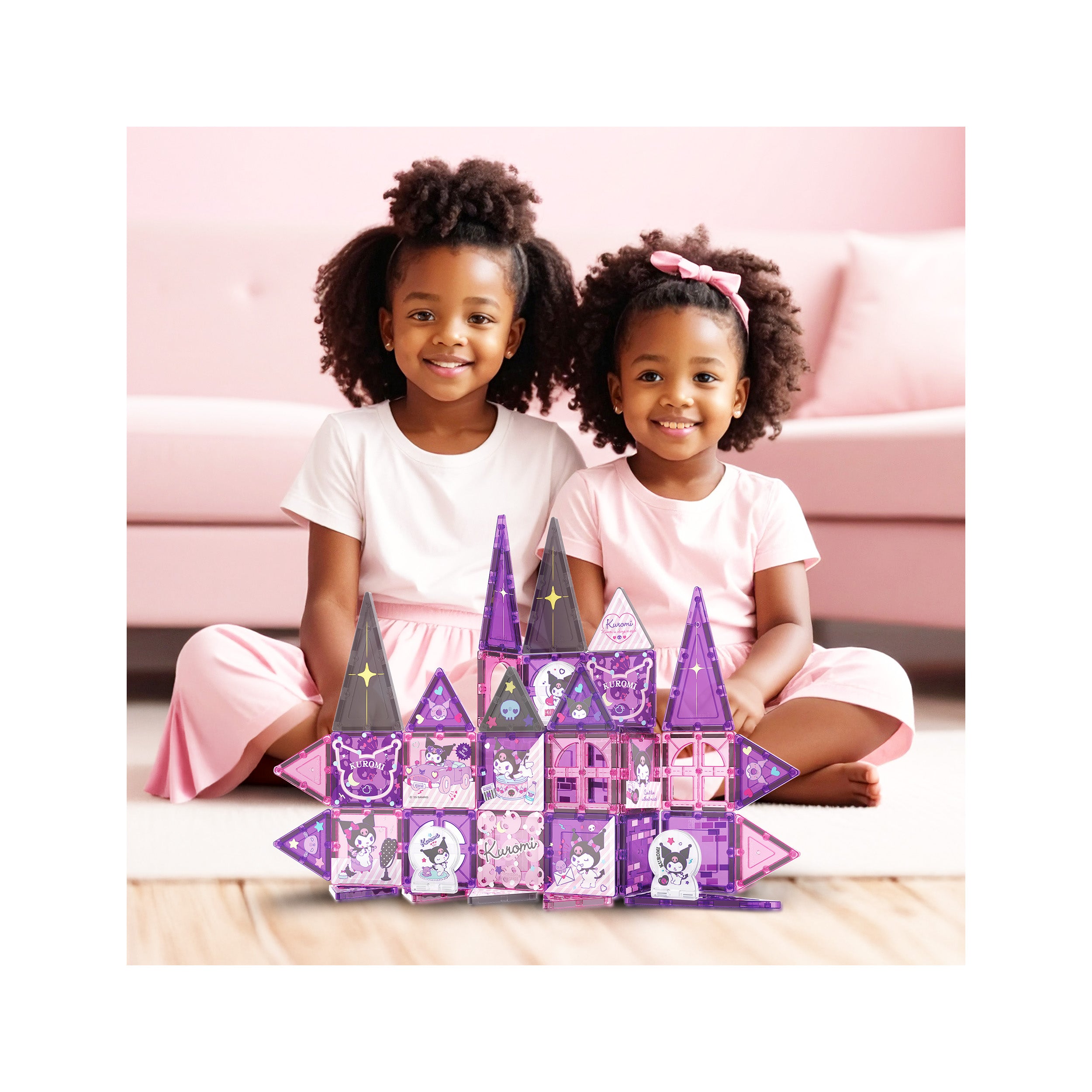 Two smiling girls with a purple and pink Kuromi magnetic tile castle on a playroom floor.