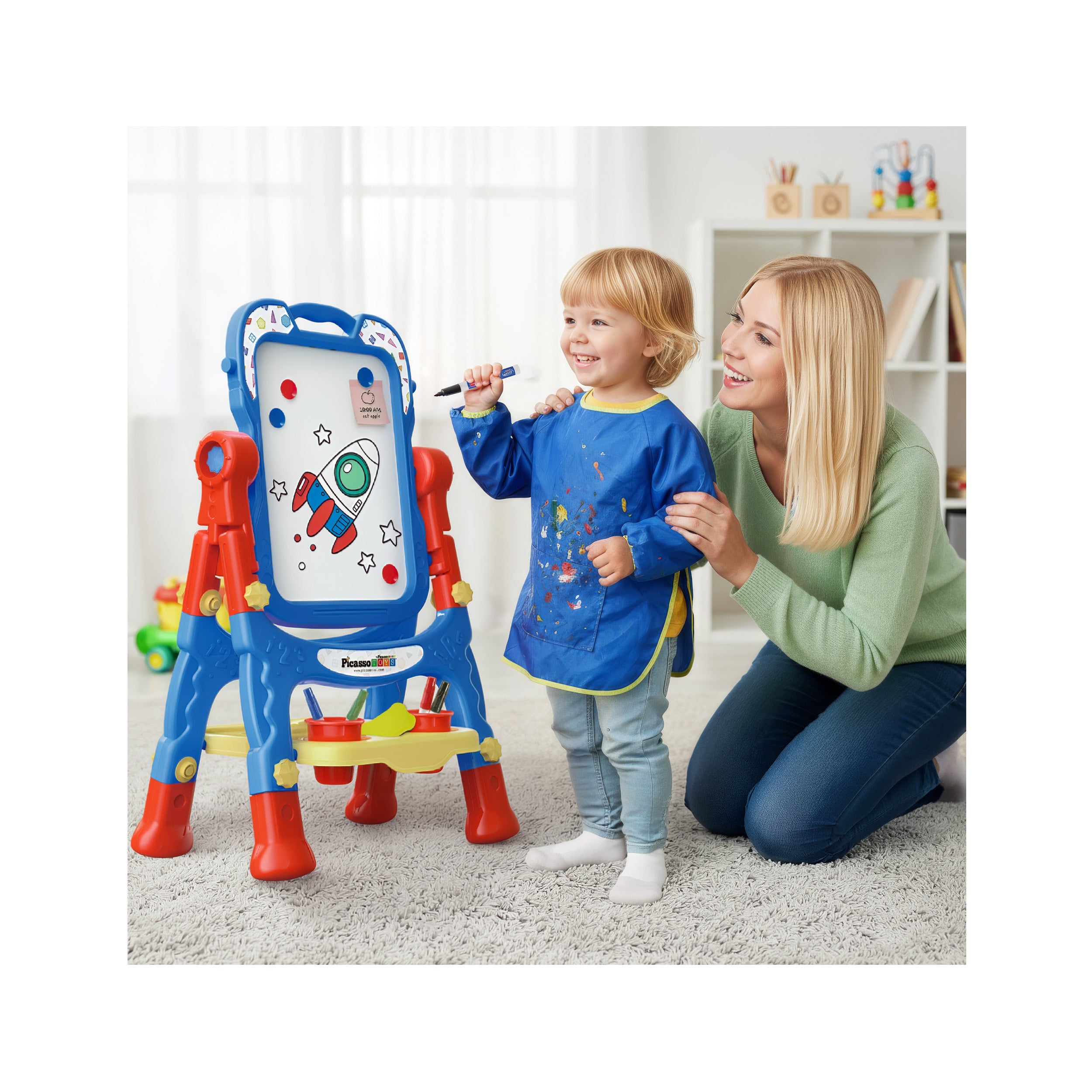 Toddler in a blue smock drawing on a colorful standing art easel while a smiling woman watches.