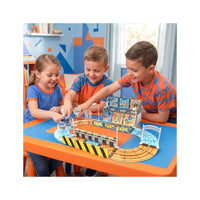 Three children are gathered around a colorful toy race track set on a table in a child's room. The toy set includes curved wooden tracks, a small bridge, a starting/finish line, and a small vehicle.