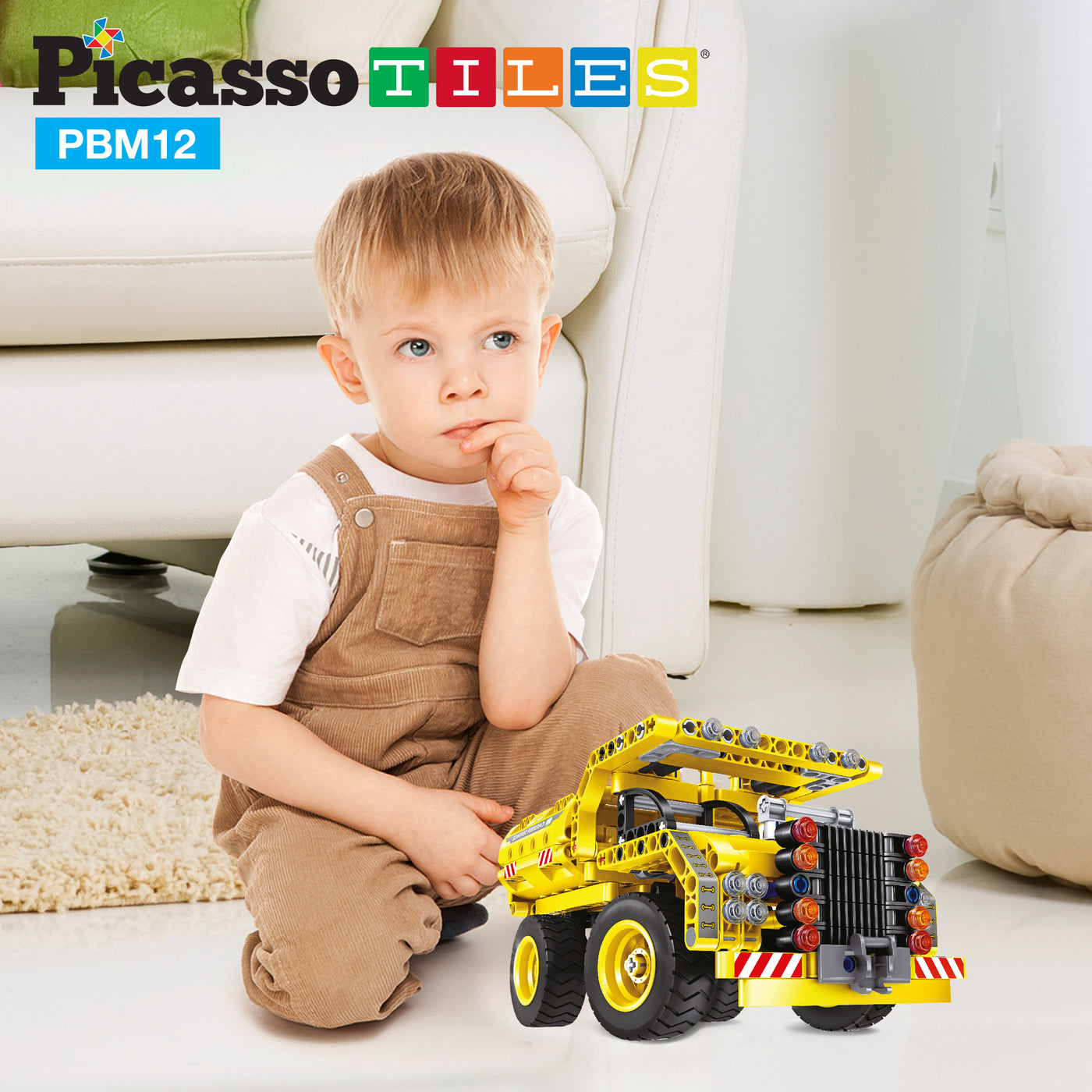 Young boy in overalls sitting on the floor next to a large yellow toy dump truck model.